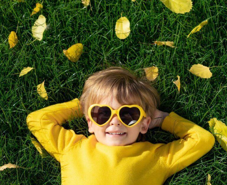Банер для блогу до статті про сонцезахисні окуляри - дитина в жовтій кофті лежить на траві, руки закинуті за голову, на ній жовті окуляри у формі сердечок