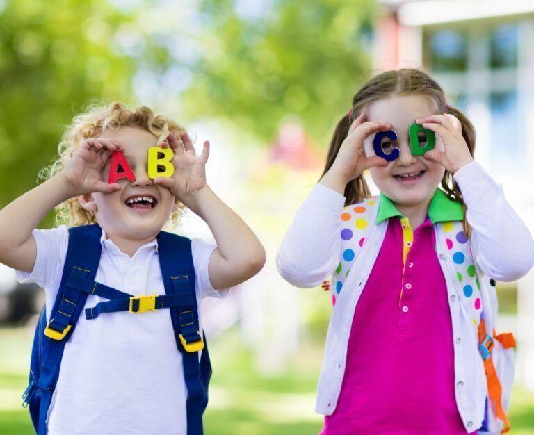 Банер до блогу до статті про підготовку до видачі довідки в школу та садок: хлопчик і дівчинка стоять з рюкзаками на фоні школи і тримають букви A, B, C, D біля очей, як окуляри