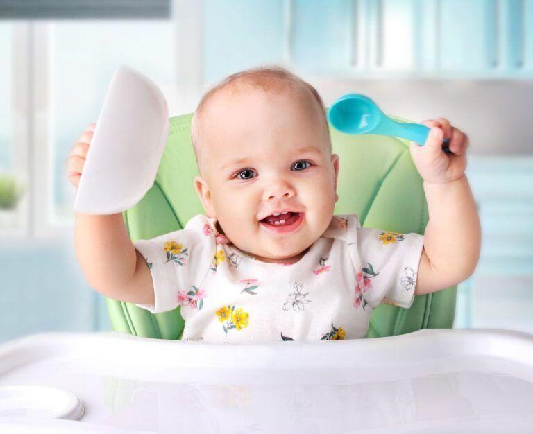 Банер для блогу до статті про харчування дітей після одного року: малюк сидить за столом, в одній руці тримає пластикову ложку, а в іншій - пластикову тарілку і посміхається