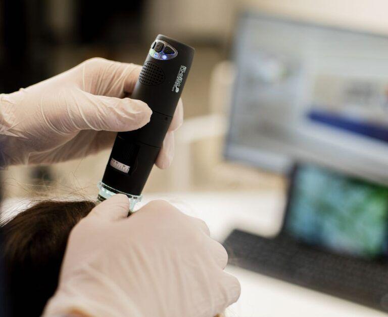 Банер для блогу до статті про підготовку до дерматологічного чекапу: лікар перевіряє стан волосся за допомогою трихоскопа в кабінеті дерматолога у Файній Клініці