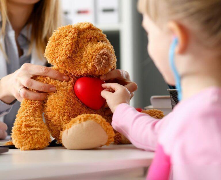 Банер для блогу до статті про підготовку до дитячого кардіологічного чекапу: дівчинка у блакитному стетоскопі слухає серце іграшкового ведмедика, якого тримає лікар