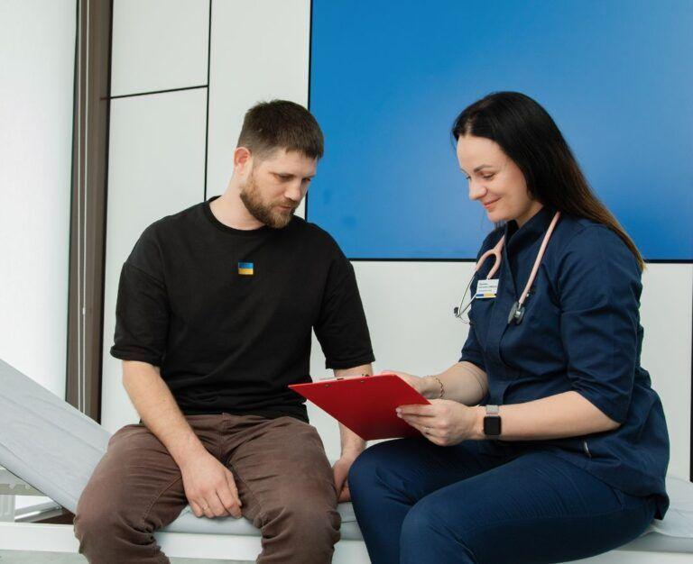 Банер до блогу для статті про підготовку до комплексного чоловічого чекапу: сімейний лікар сидить на кушетці з чоловіком-пацієнтом і проводить консультацію дивлячись в планшет в кабінеті Файної Клініки